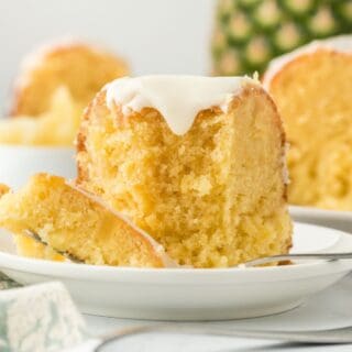 A slice of pineapple pound cake on a white plate with a fork, showing its moist interior and glaze, with a whole pineapple in the background