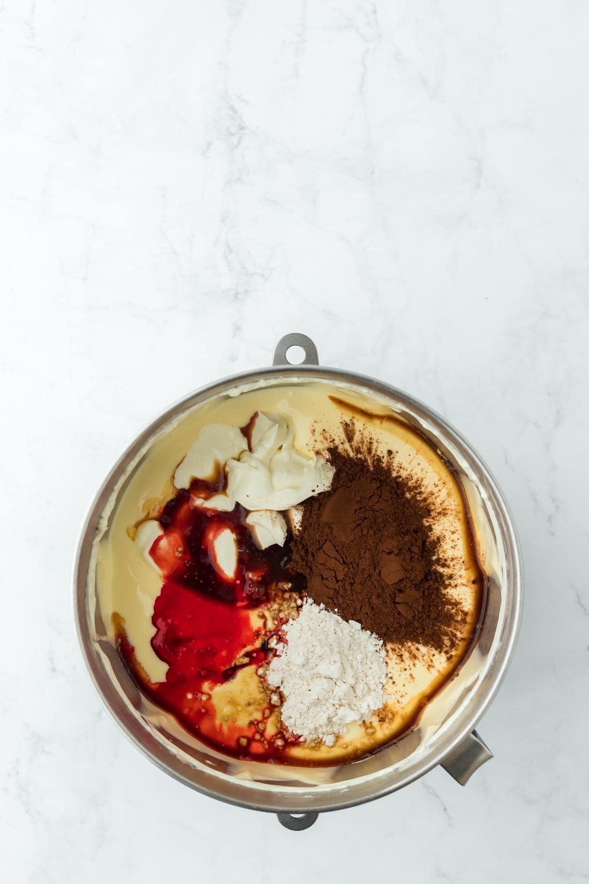 Mixing bowl with cheesecake batter, cocoa powder, flour, sour cream, vanilla, and red food coloring added on top