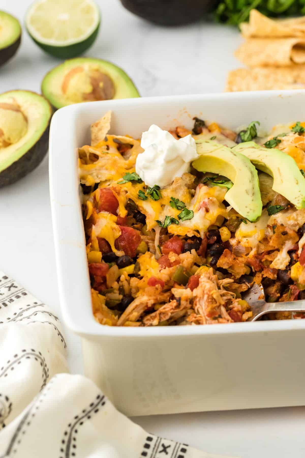 A white casserole dish filled with Southwestern chicken casserole, featuring layers of rice, beans, corn, shredded chicken, and melted cheese, garnished with avocado slices and sour cream. In the background there's avocados, tortilla chips and lime