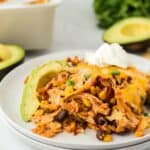 A white casserole dish filled with Southwestern chicken casserole, featuring layers of rice, beans, corn, shredded chicken, and melted cheese, garnished with avocado slices and sour cream. In the background there's avocados, tortilla chips and lime