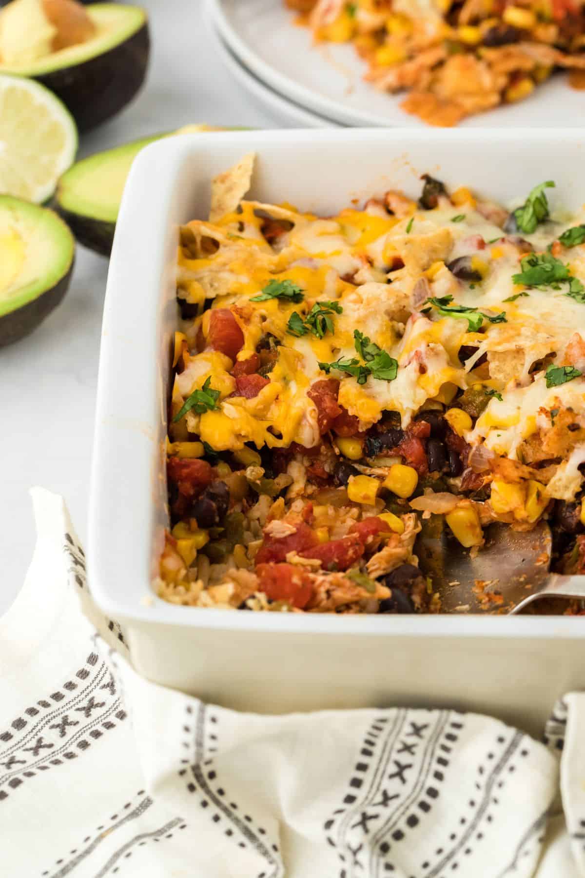 A white casserole dish filled with Southwestern chicken casserole, featuring layers of rice, beans, corn, shredded chicken, and melted cheese, garnished with chopped cilantro. Next to it are avocados and lime, with more of the casserole plated in the background