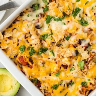 Overhead shot of a white rectangular baking dish filled with Southwestern chicken casserole, topped with melted cheese and chopped fresh cilantro, with avocados next to it