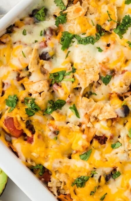 Overhead shot of a white rectangular baking dish filled with Southwestern chicken casserole, topped with melted cheese and chopped fresh cilantro, with avocados next to it