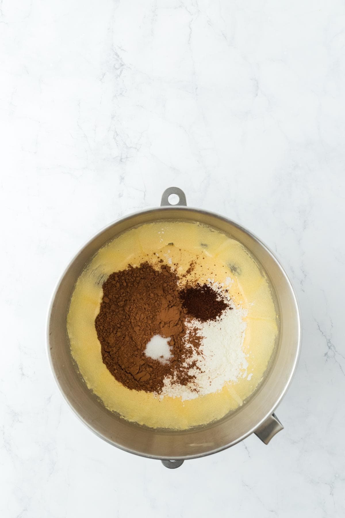 Dry ingredients including cocoa powder and coffee added to a creamy egg mixture in a mixing bowl