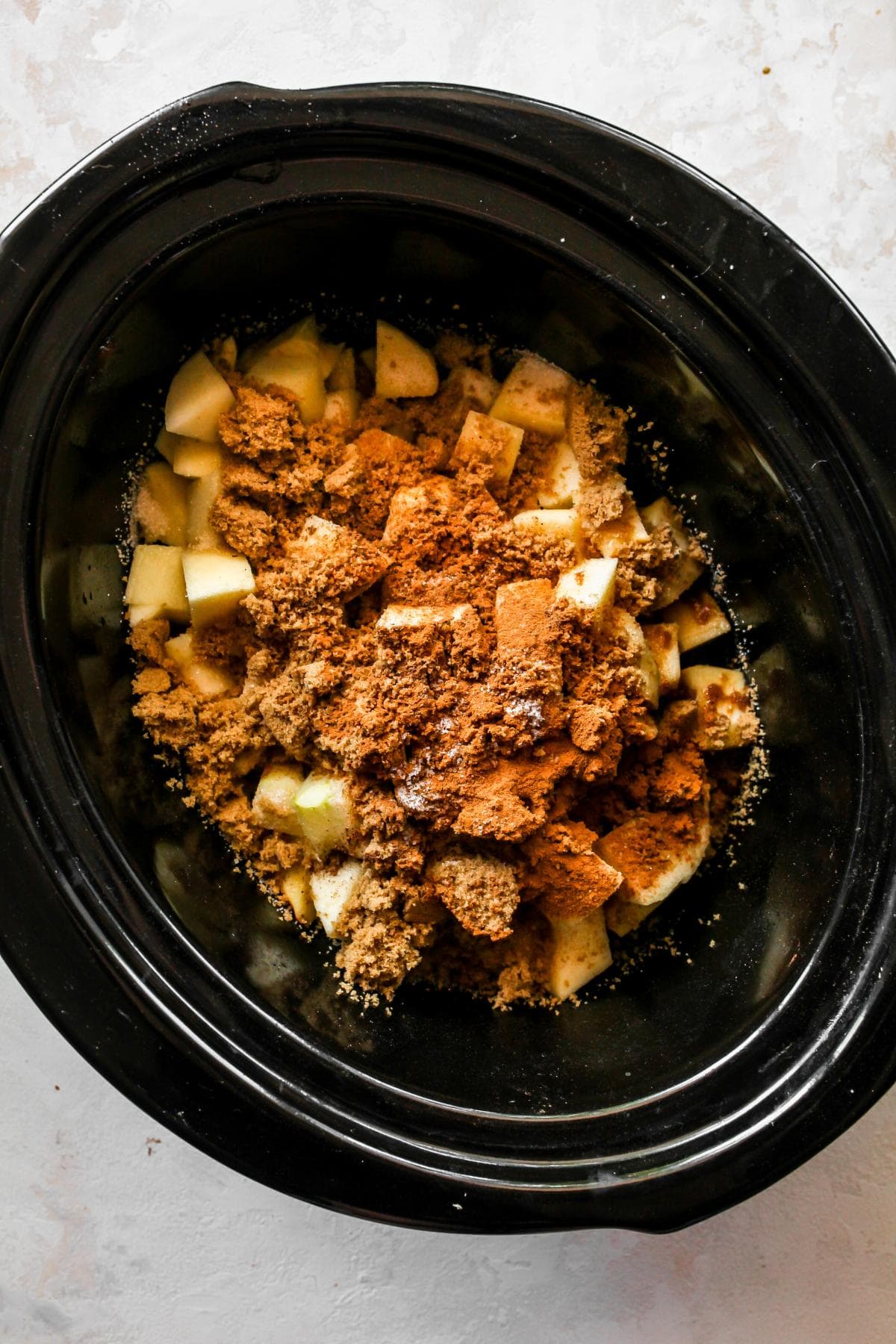 Diced apples are in a black slow cooker, covered with brown sugar and cinnamon