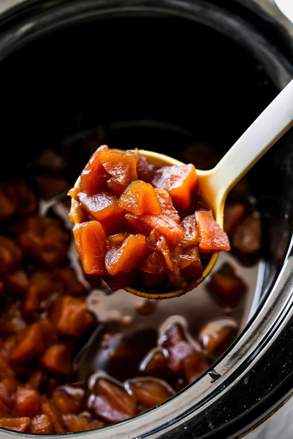 A spoon holds a scoop of cooked, diced apples in a reddish-brown sauce, lifted from a slow cooker