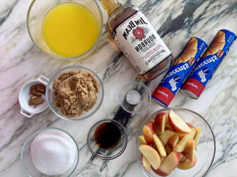 An ingredient shot over a marbled countertop with apples in a bowl along with crescent roll dough, whisky, brown sugar, butter and other ingredients