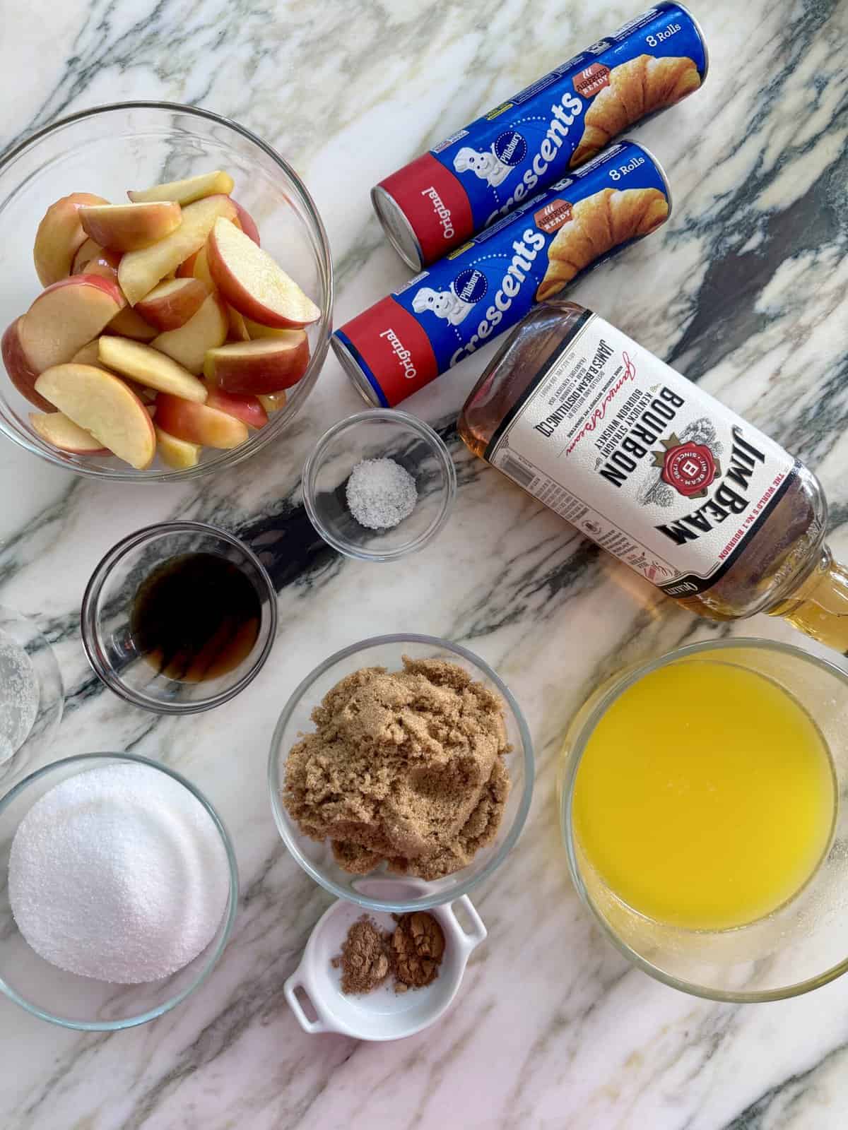 An ingredient shot over a marbled countertop with apples in a bowl along with crescent roll dough, whisky, brown sugar, butter and other ingredients