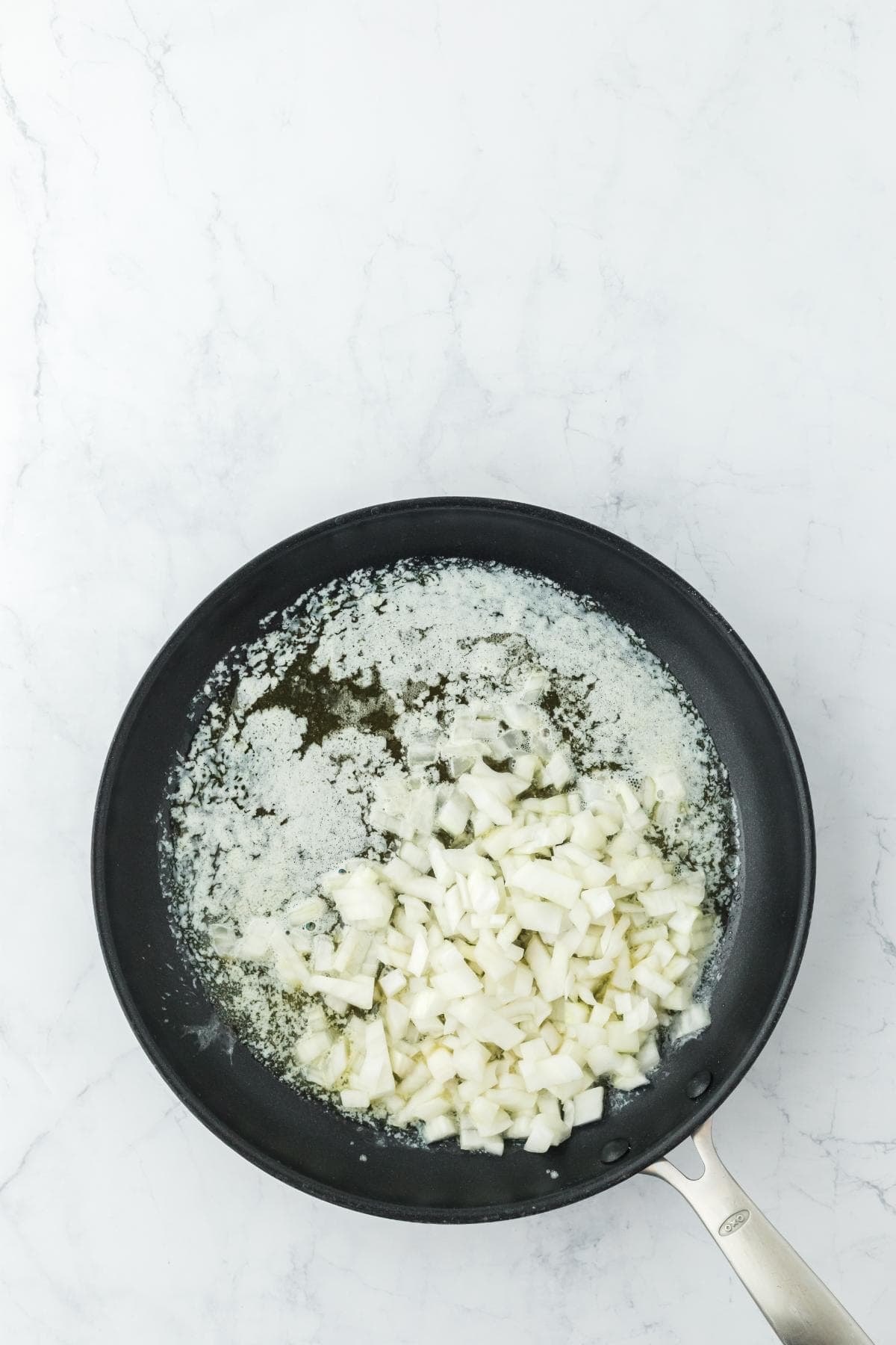A black frying pan on a white marble surface with melted butter and diced white onions