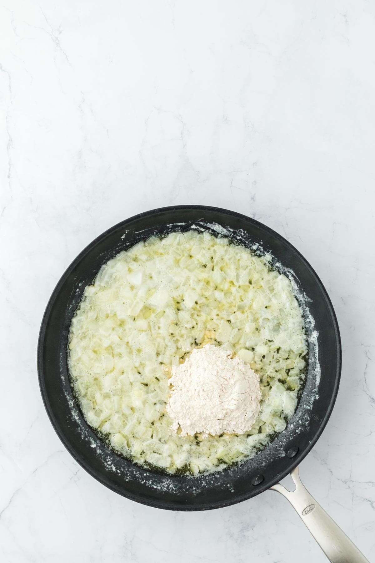 A black frying pan on a white marble surface containing sautéed diced onions, with a pile of white flour and a small amount of dried herbs added