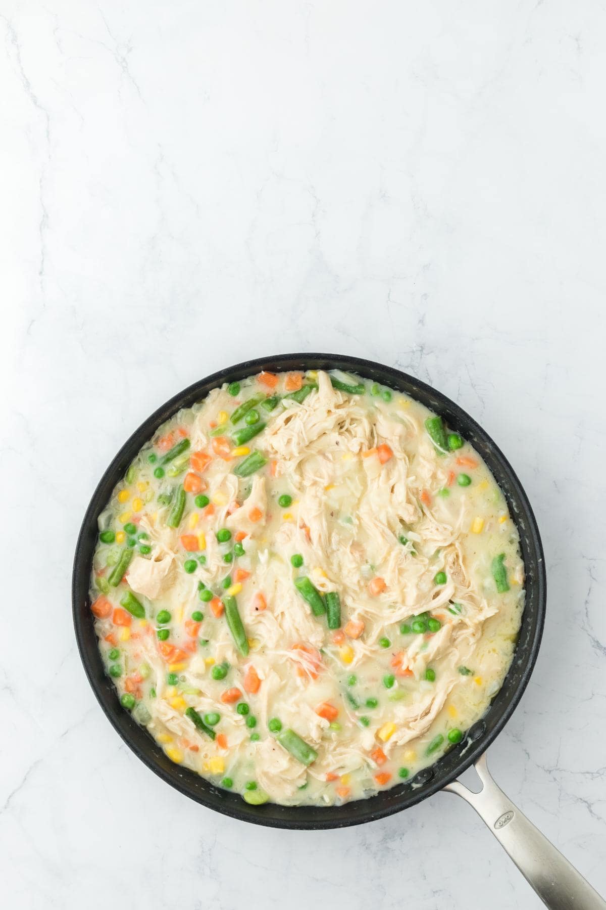A black frying pan on a white marble surface containing a creamy chicken pot pie filling with mixed vegetables and shredded chicken, stirred together