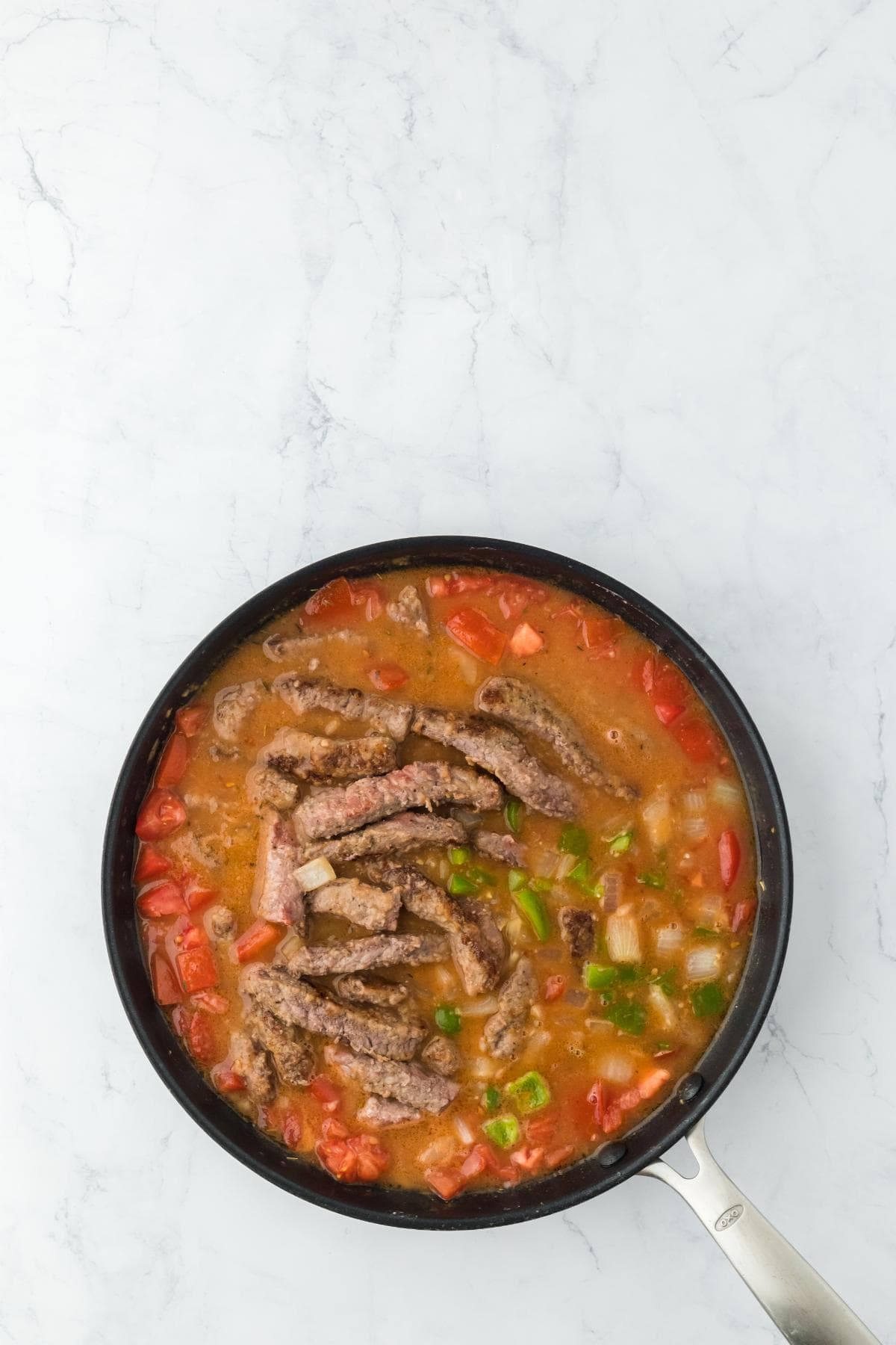 A black frying pan on a white marble surface containing browned meat strips added to a simmering mixture of diced tomatoes, onions, and green bell peppers in a reddish sauce
