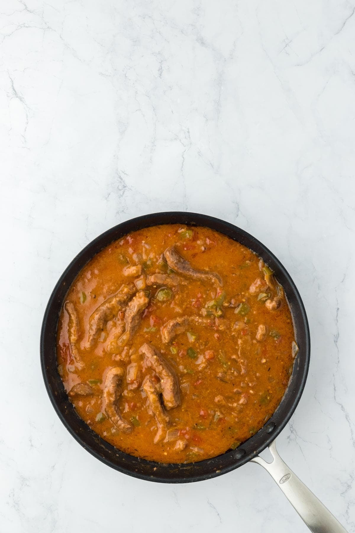A black frying pan on a white marble surface filled with a rich, reddish-brown stew containing strips of meat, diced tomatoes, onions, and green bell peppers
