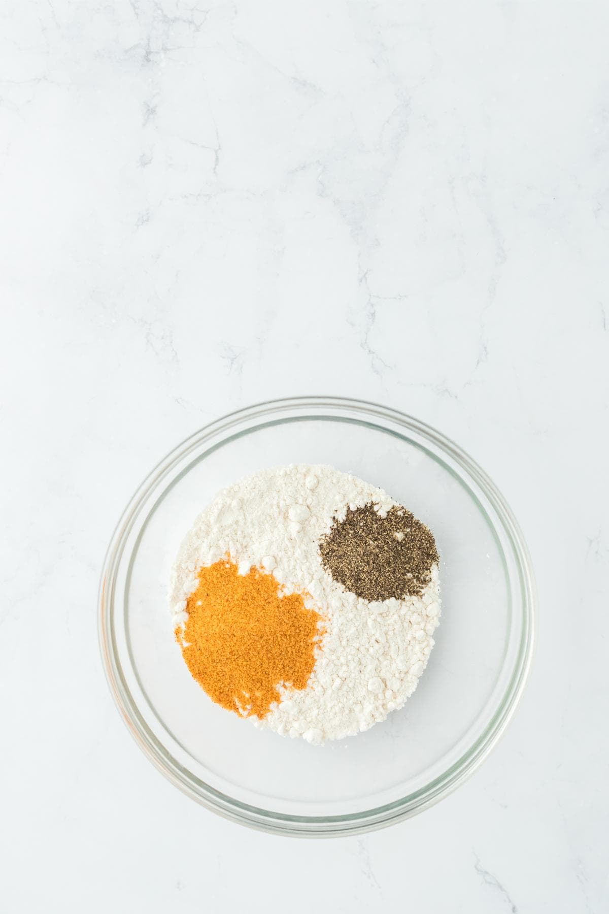 A clear glass bowl on a marble counter with white flour, a pile of spices, and a pile of black pepper
