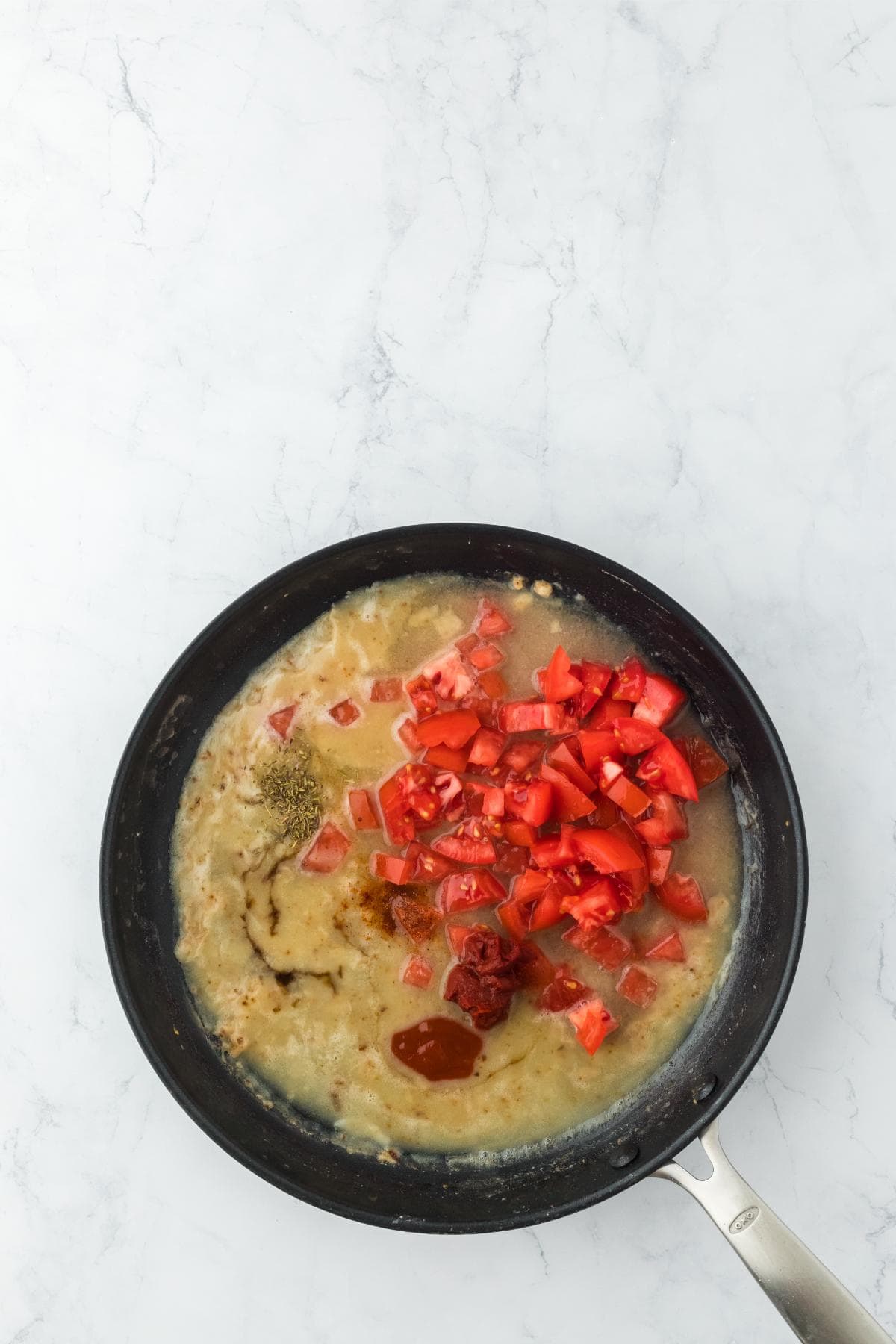A black frying pan on a white marble surface containing a simmering liquid with diced red tomatoes, a blob of red paste, and dark seasonings