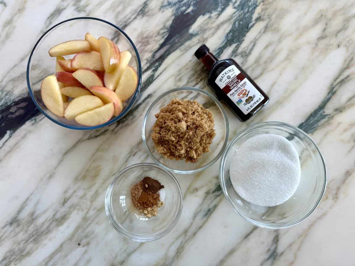 Slices of apples, brown sugar, granulated sugar, vanilla, spices and salt in clear bowls against a marble background