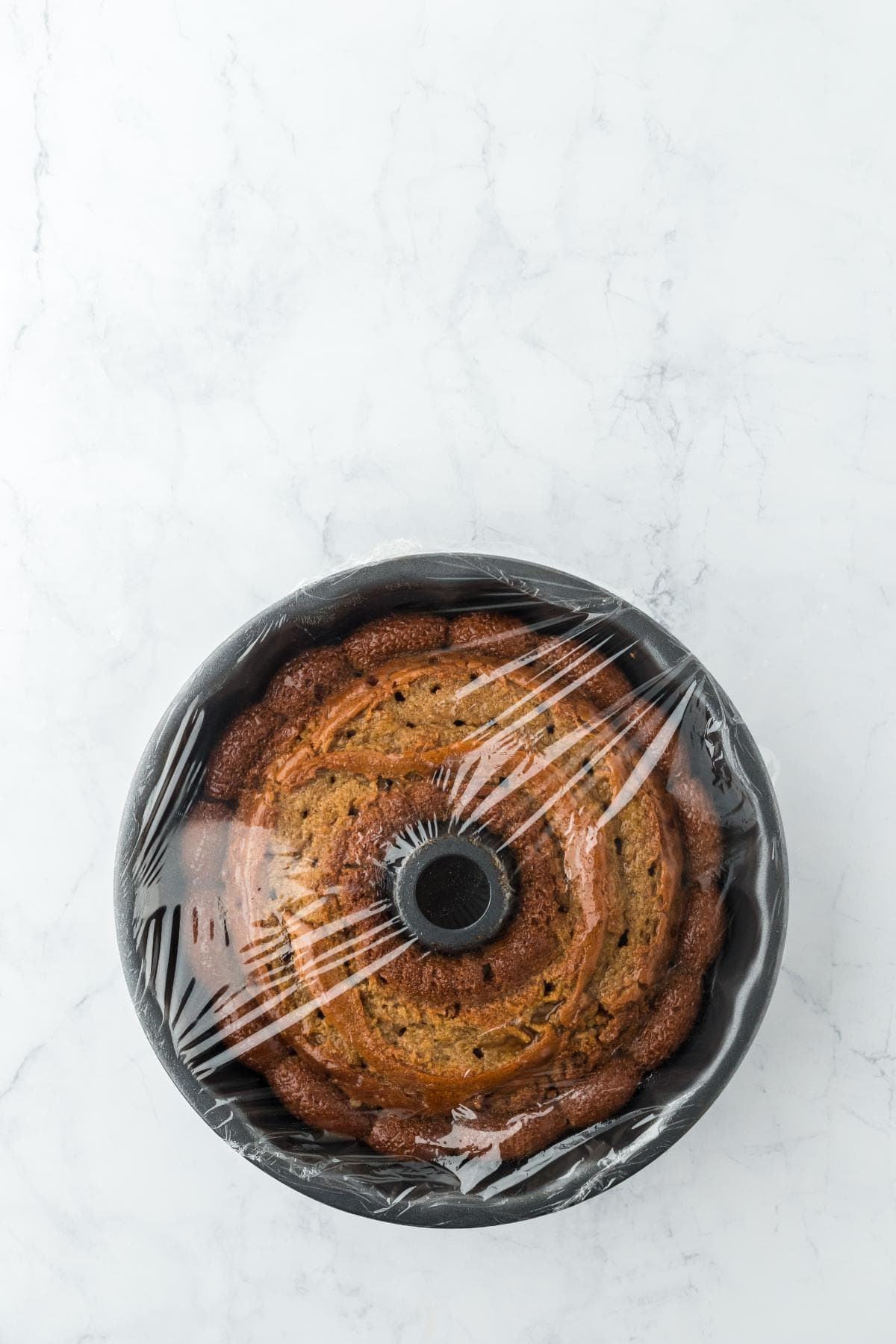 A freshly baked bundt cake with holes poked in cooling in its bundt pan, covered with clear plastic wrap