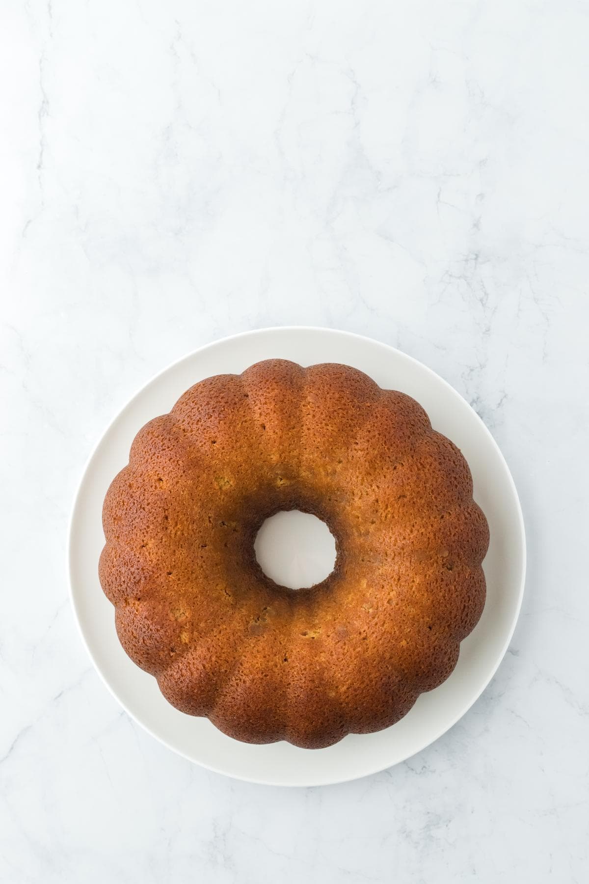 A baked rum cake is resting on a white round plate, centered on a white marble surface