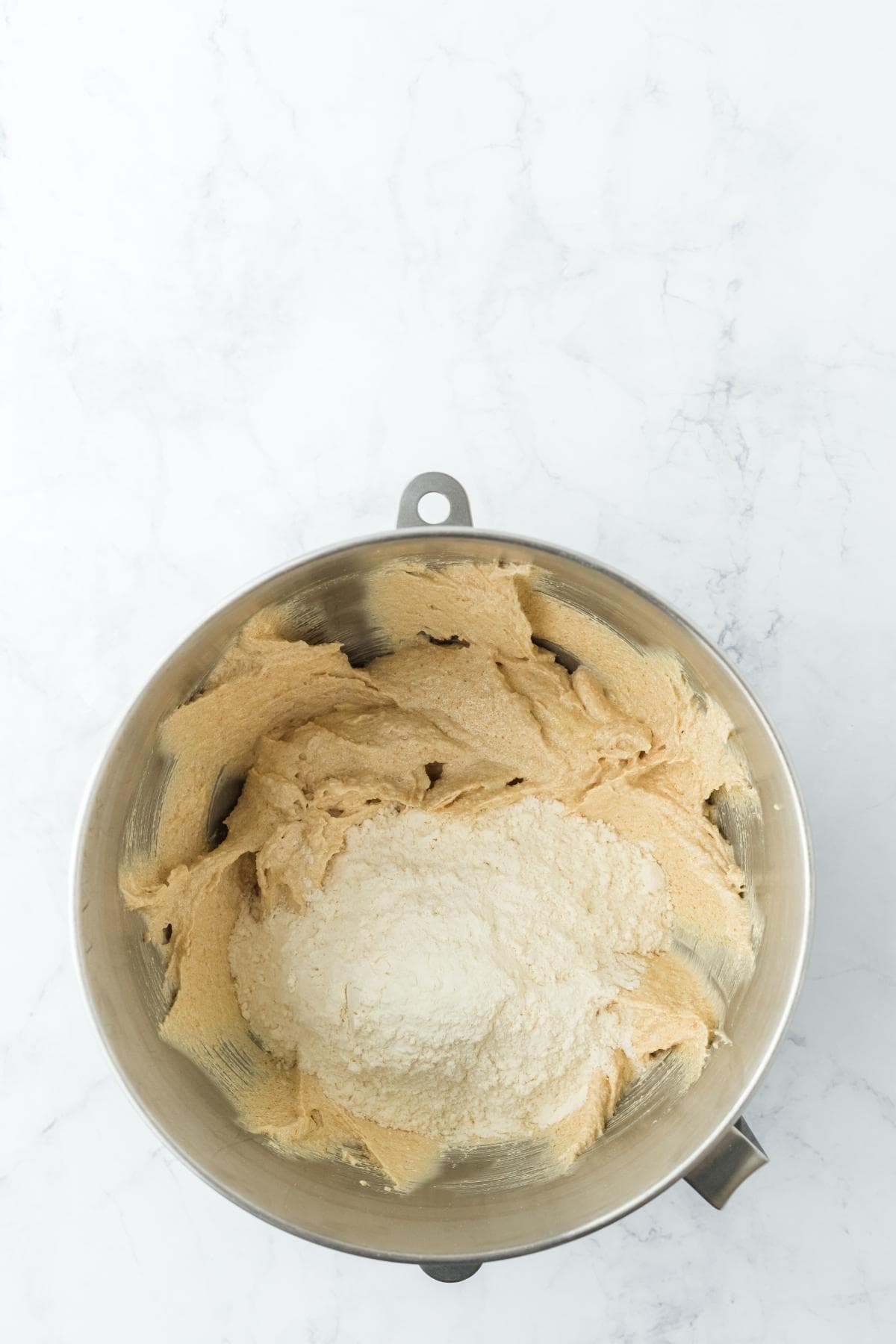 A stainless steel mixing bowl on a marble counter contains light brown cake batter with a pile of white flour on top
