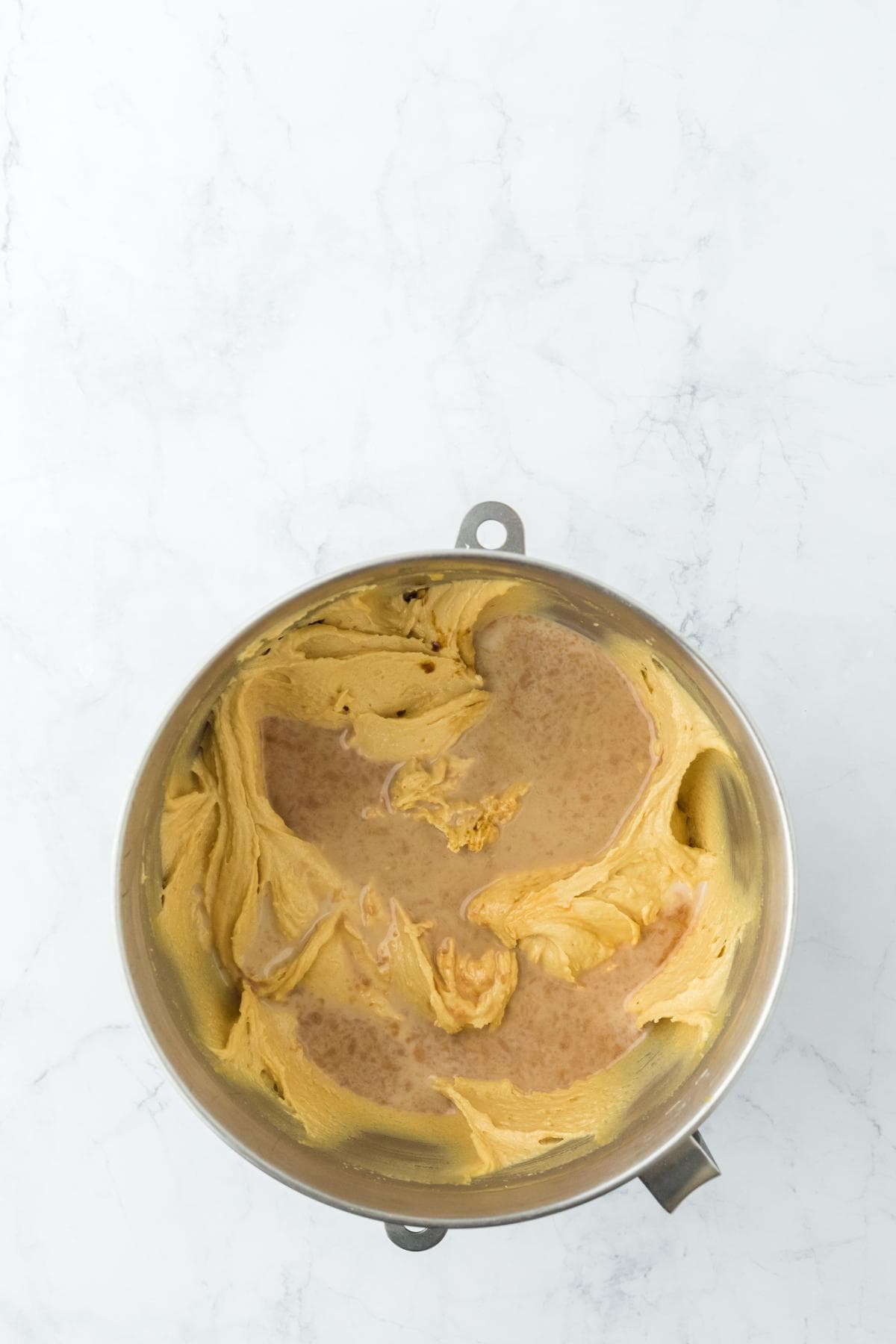 A stainless steel mixing bowl on a marble counter contains light brown cake batter mixed with the liquid ingredients