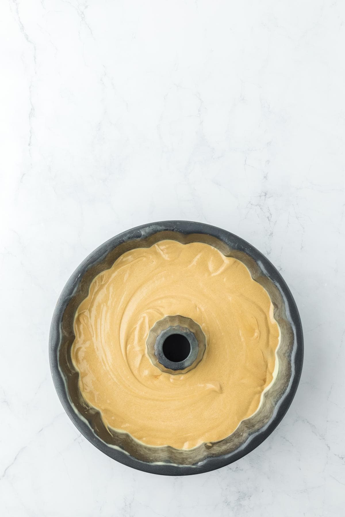 A dark gray fluted bundt pan on a white marble surface contains light brown cake batter, ready for baking
