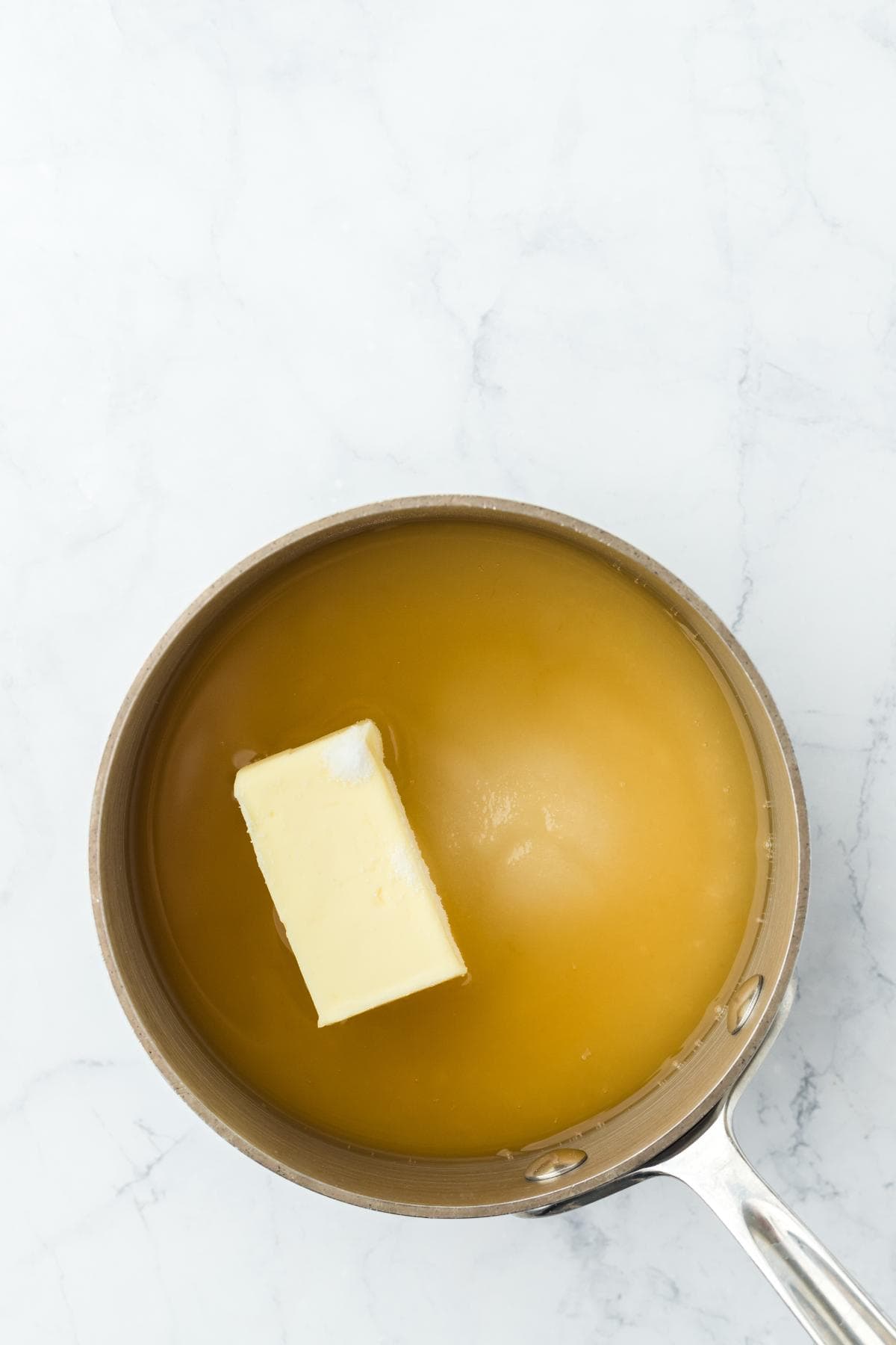 A metal saucepan on a white marble counter holds a yellow liquid with a stick of butter floating in it