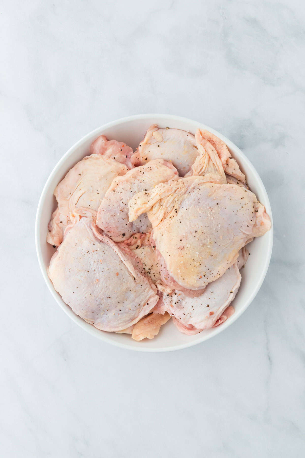 Bone in skin on chicken in a white bowl seasoned on a white background