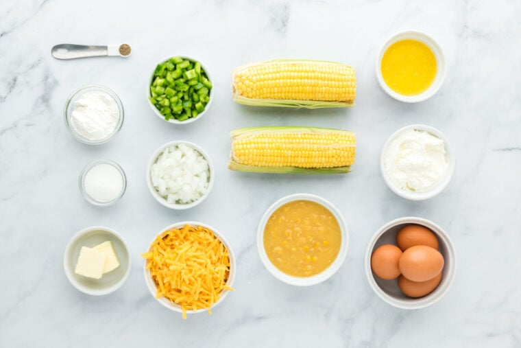 Corn on the cob, sugar, eggs, flour, cheese, veggies in white bowls on a white background