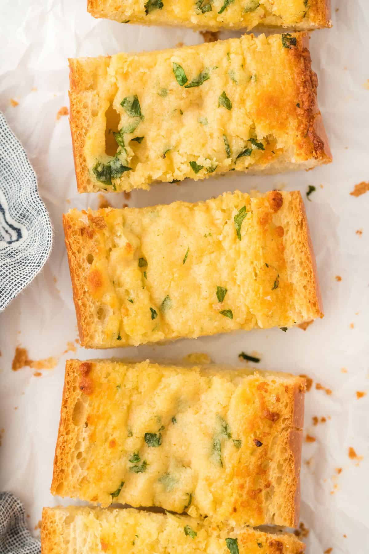 Slices of garlic bread, golden brown and topped with garlic and parsley, laid on a white parchment paper