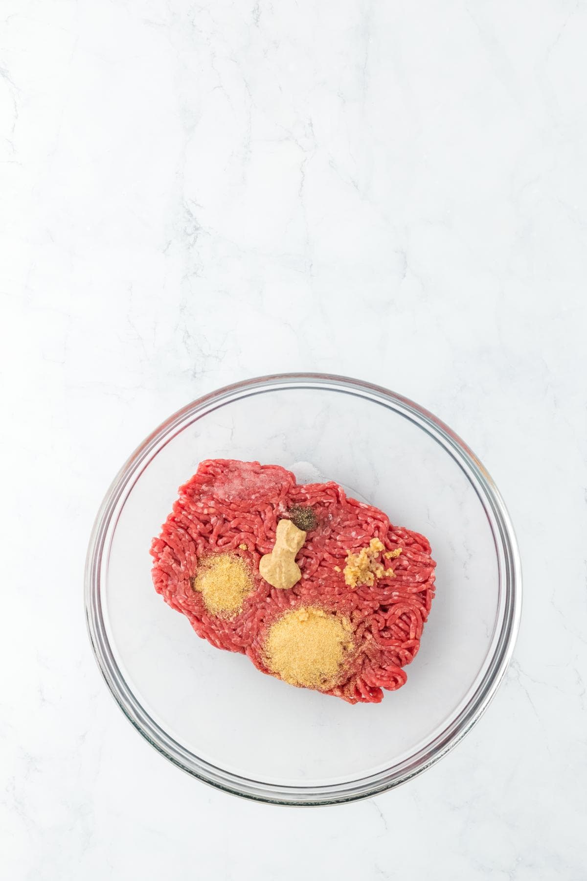 A clear glass bowl on a white marble surface with raw ground beef mixed with spices, black pepper, and a small dollop of dijon mustard