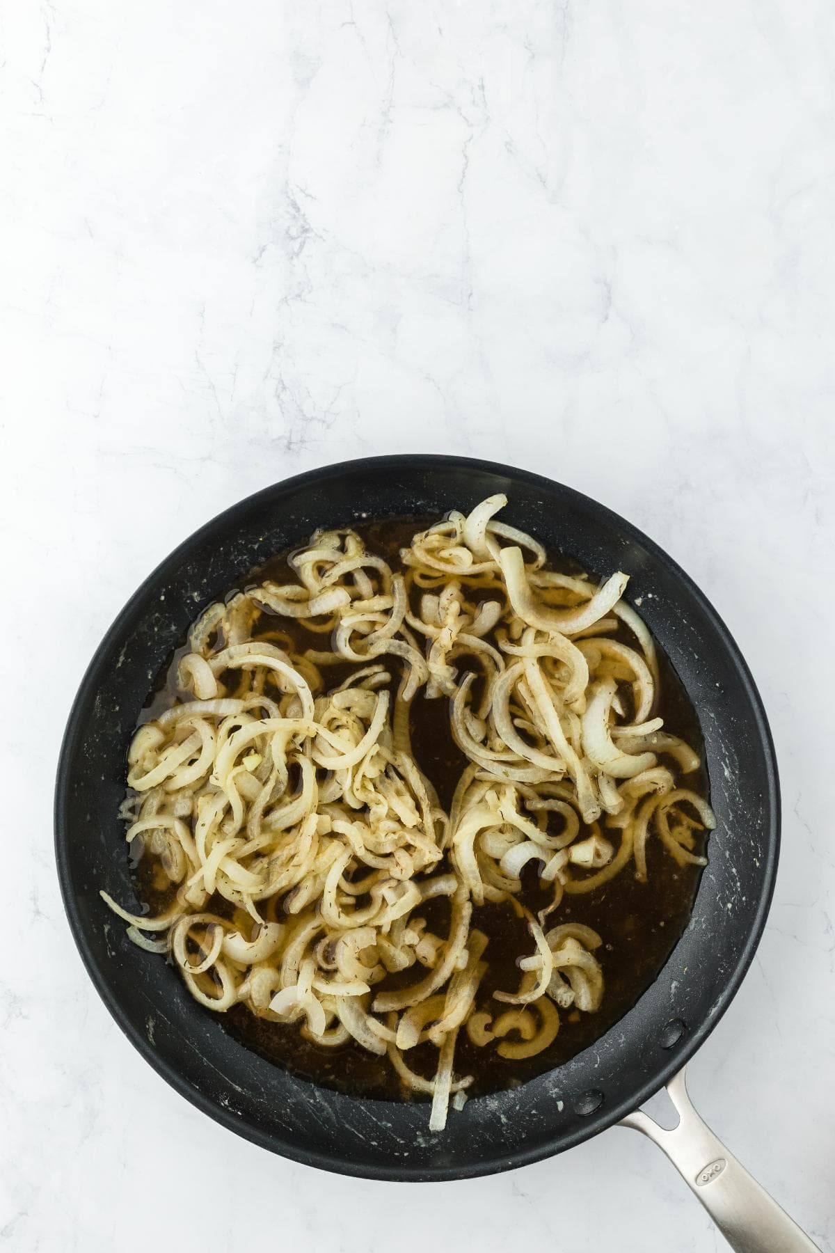 Sliced onions simmering in a dark liquid in a black frying pan on a white marble surface