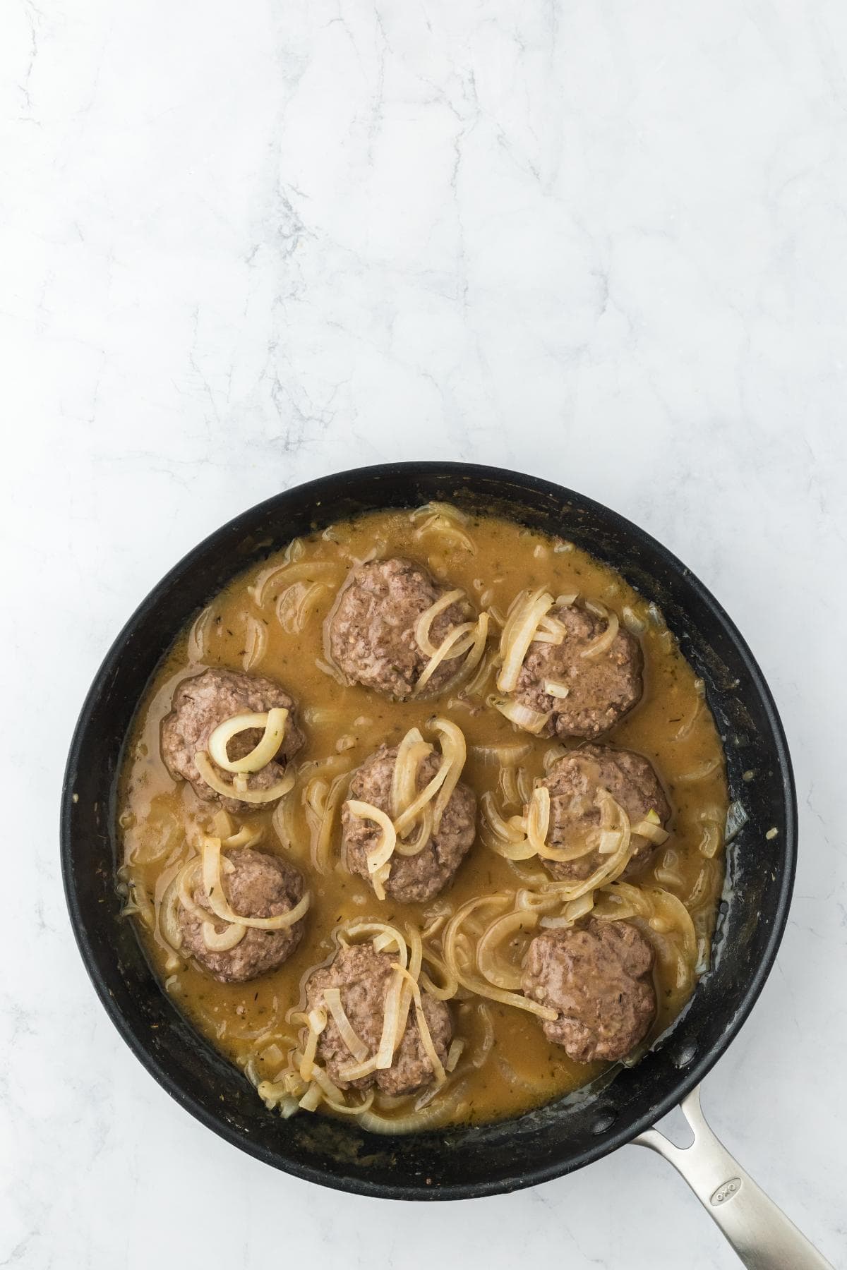 Seven cooked hamburger patties submerged in a thick, savory brown gravy with sliced onions in a black frying pan on a white marble surface