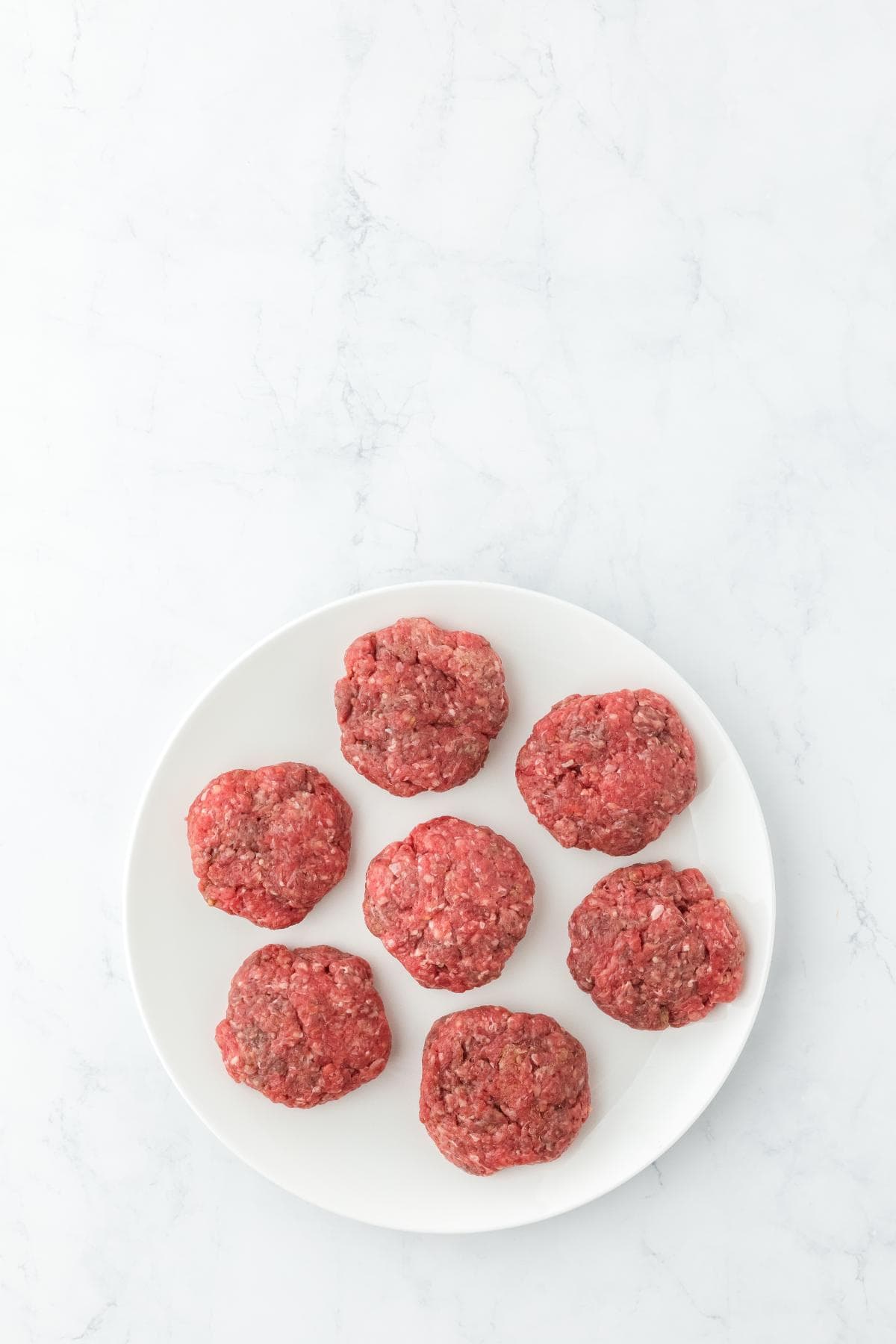 A white plate on a white marble surface with seven raw, round hamburger patties