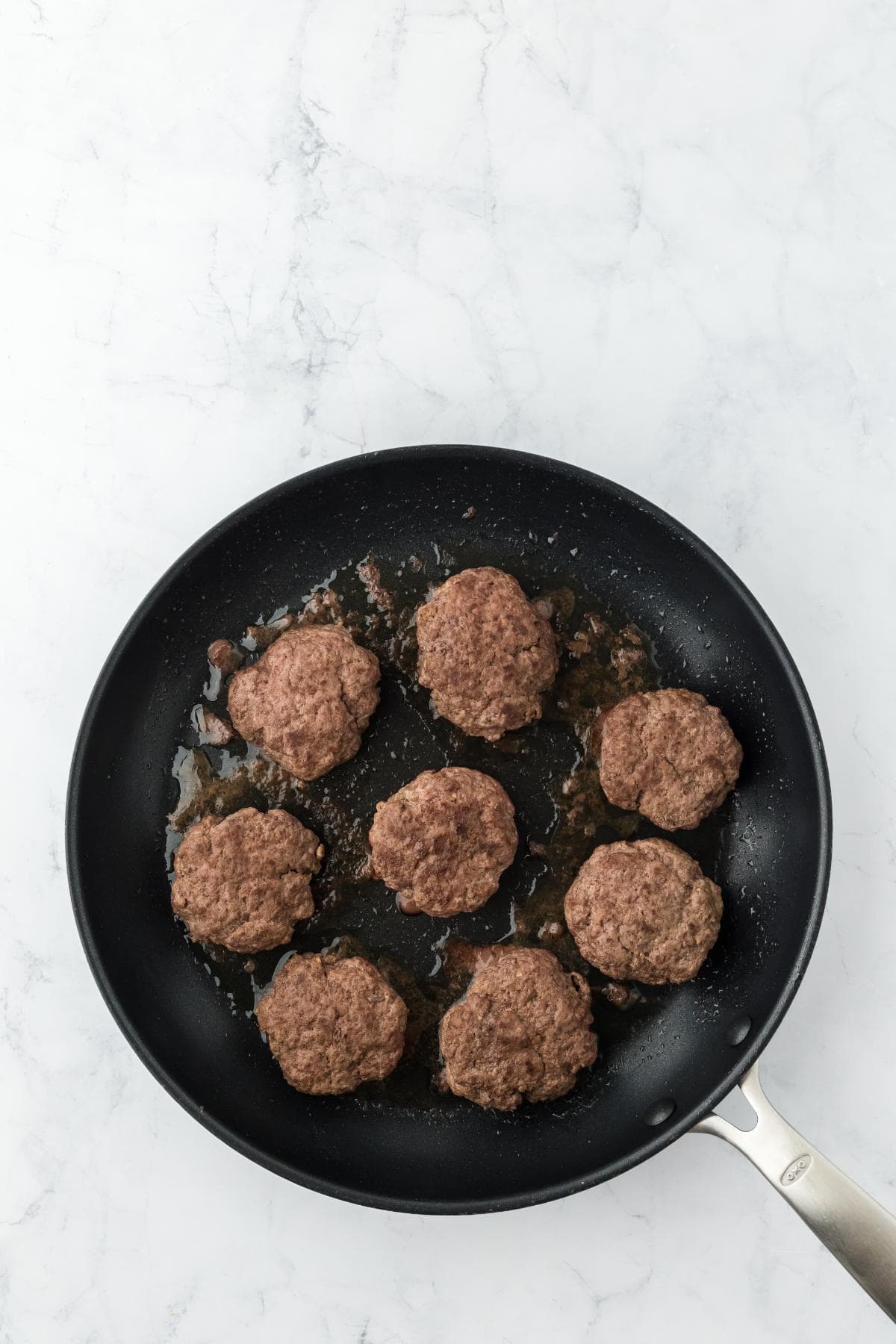 Seven browned hamburger patties cooking in a black frying pan with some rendered fat, on a white marble surface