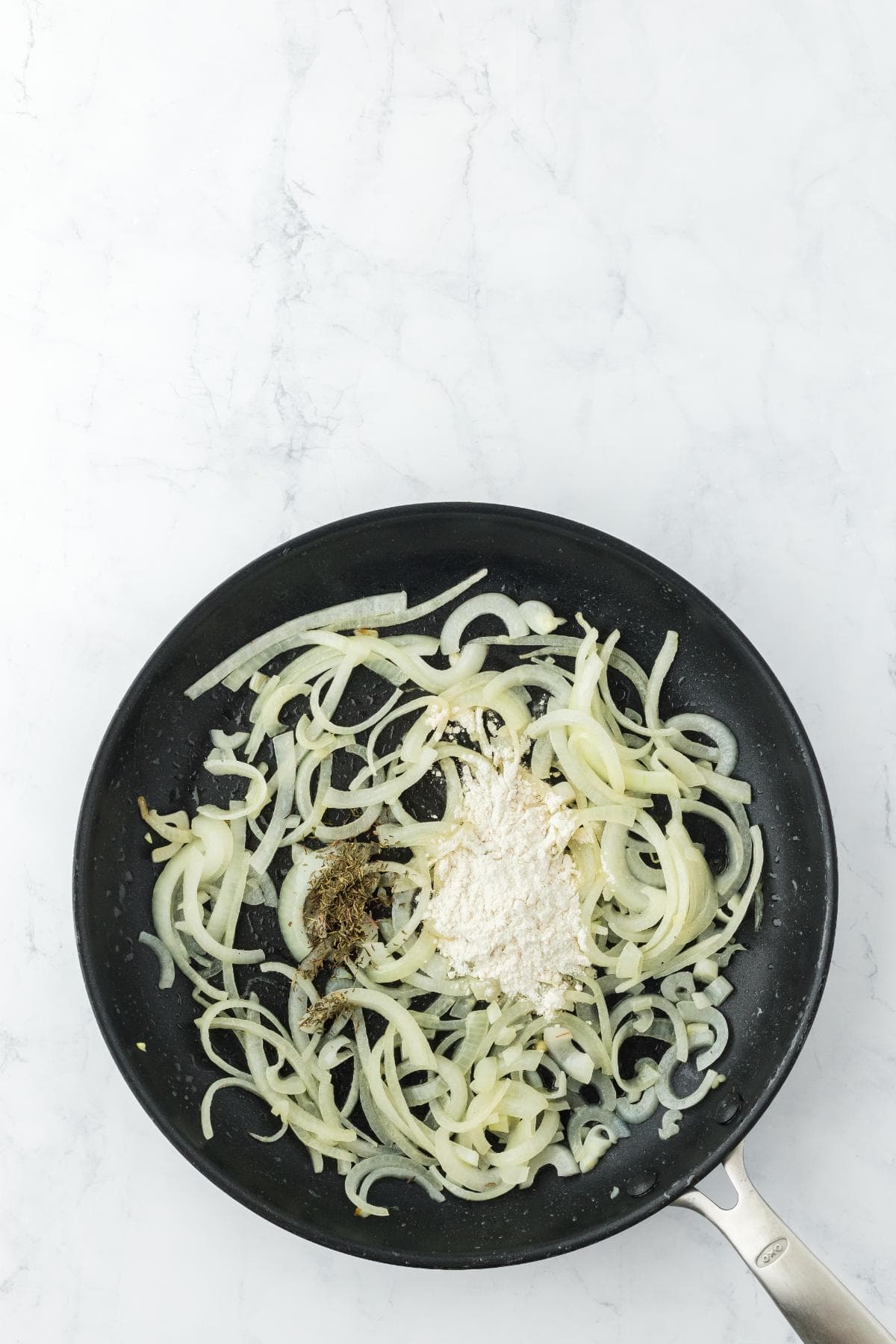 A black frying pan on a white marble surface with sautéed sliced onions and a pile of white flour and spices