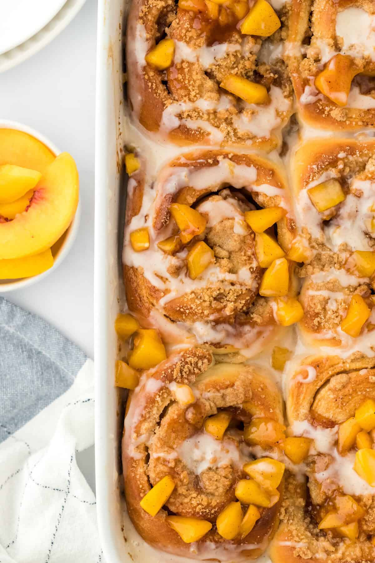 Peach cinnamon rolls in a white baking dish on a white background with a bowl of peach slices ready to enjoy