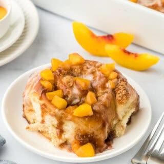 A single peach cobbler cinnamon roll on a white plate in a white background with fresh peach slices in the background with cinnamon rolls in the background