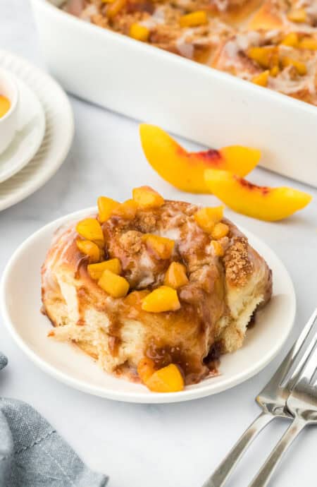 A single peach cobbler cinnamon roll on a white plate in a white background with fresh peach slices in the background with cinnamon rolls in the background