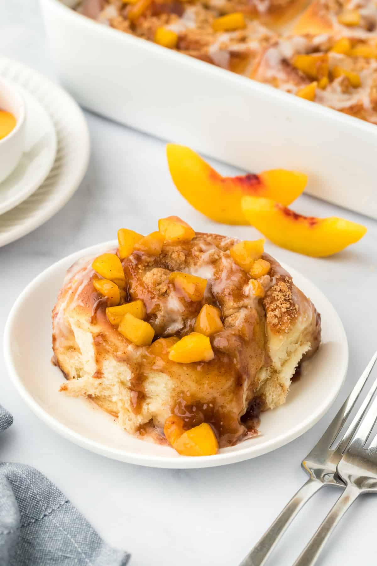 A single peach cobbler cinnamon roll on a white plate in a white background with fresh peach slices in the background with cinnamon rolls in the background