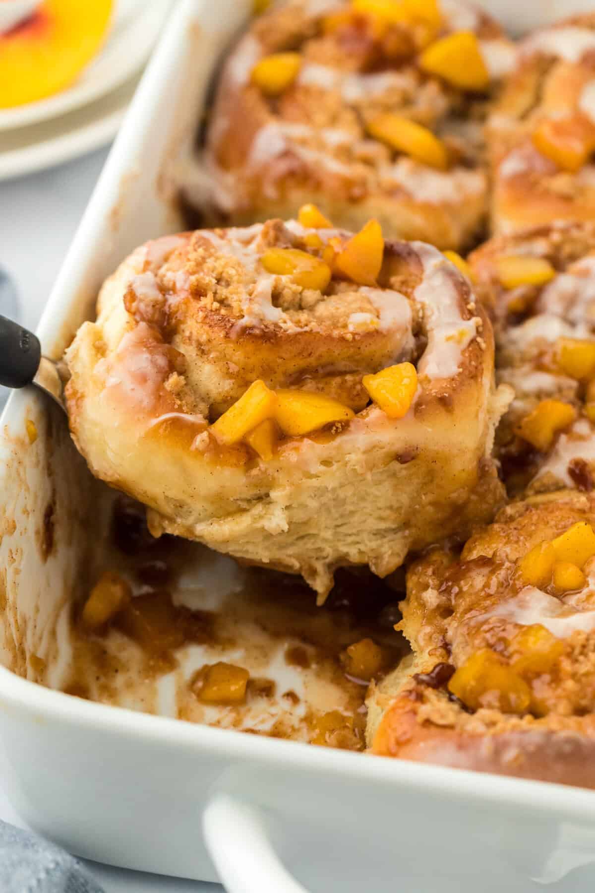 A peach cobbler cinnamon roll being lifted out of the white baking pan with a spatula