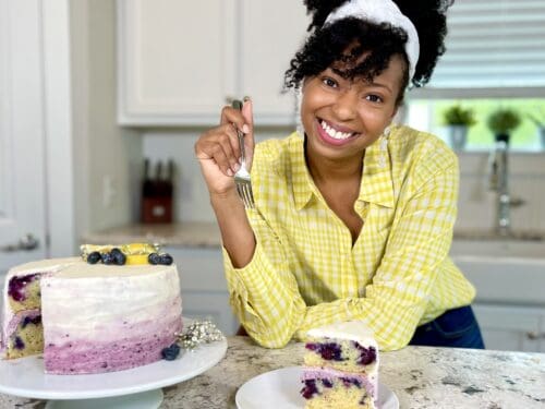 Jocelyn Delk Adams with a slice of lemon blueberry cake on a plate next to whole layer cake in a kitchen