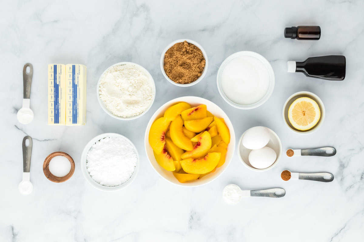 Peaches, sugars, flour, butter and spices in white bowls on a white background