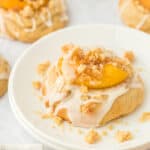 A peach cobbler cookie sitting on a white plate with extra icing surrounded with other peach cookies on a white background