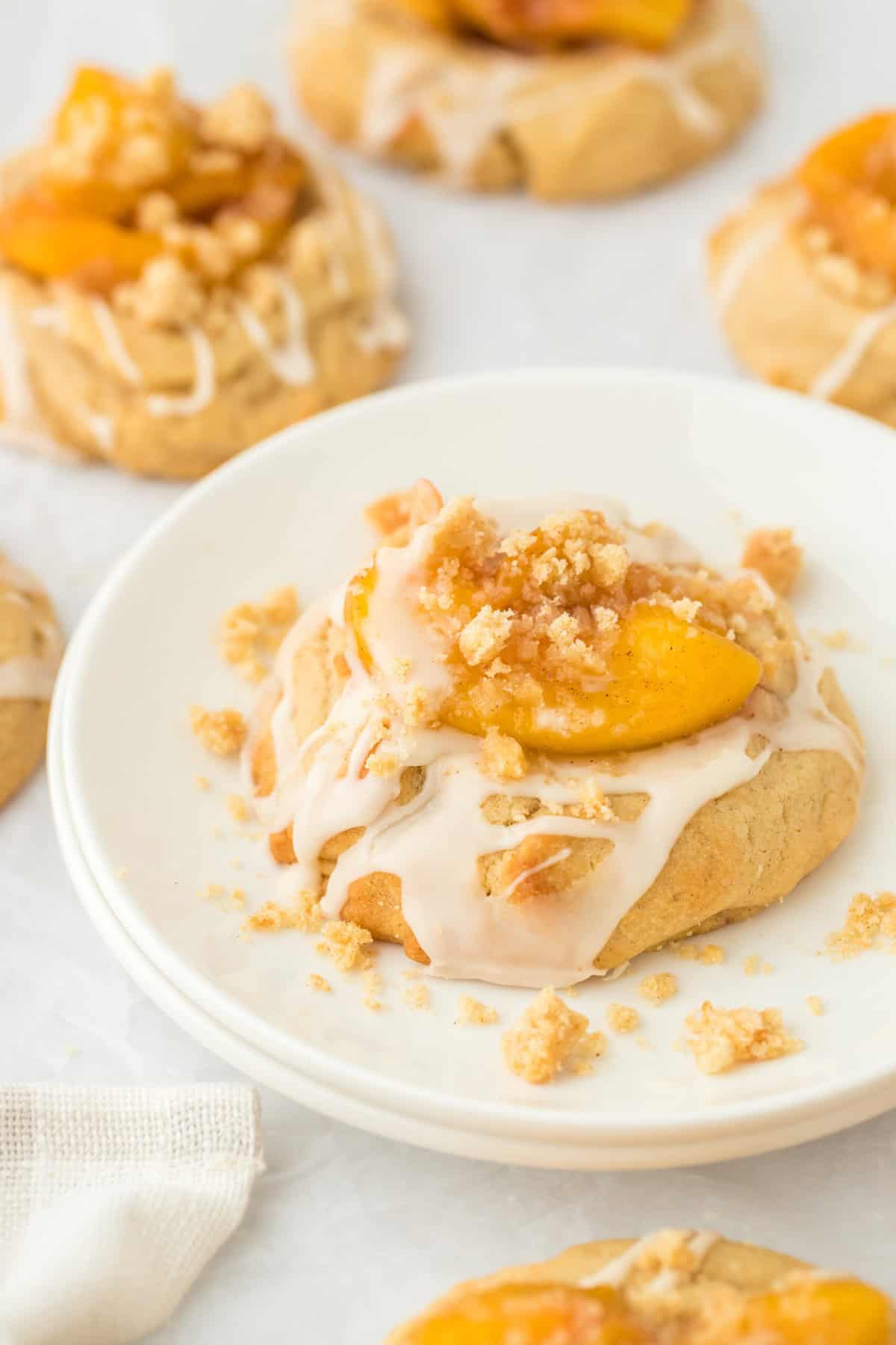 A peach cobbler cookie sitting on a white plate with extra icing surrounded with other peach cookies on a white background