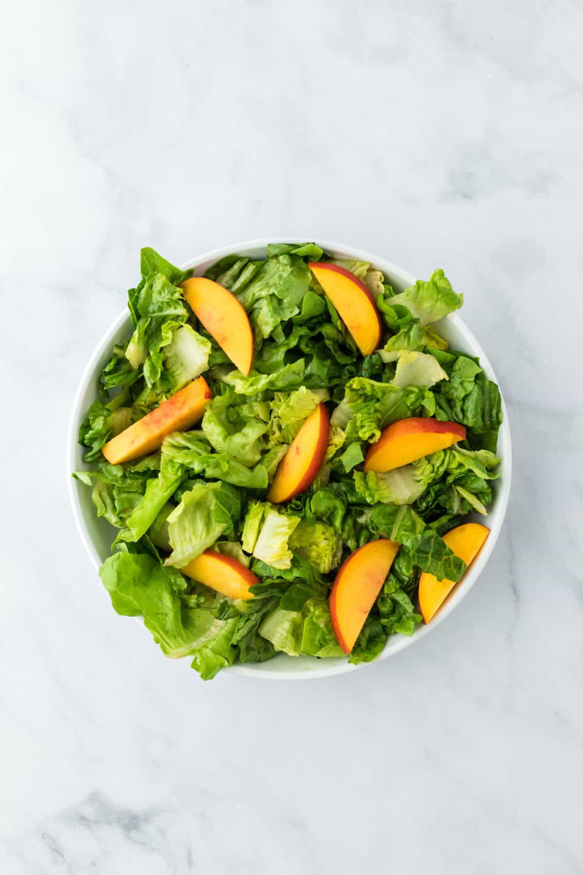 Sliced ripe peaches added to lettuce in a white serving bowl on a white background