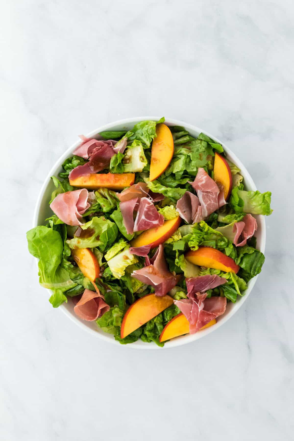 Prosciutto added to a peach salad recipe in a white bowl on a white background