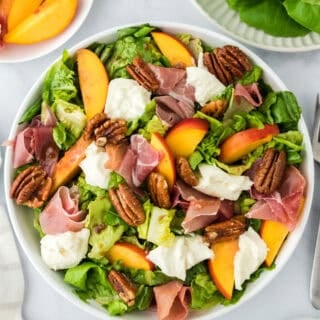 A peach salad with burrata and prosciutto in a white bowl with lettuce on the side along with fresh peaches on a white background