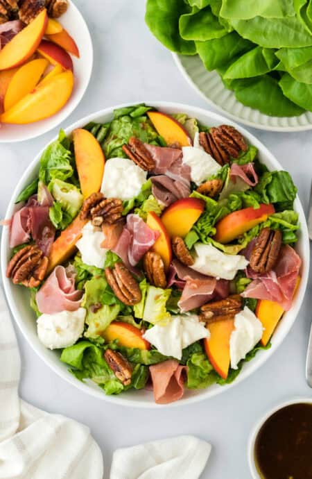 A peach salad with burrata and prosciutto in a white bowl with lettuce on the side along with fresh peaches on a white background