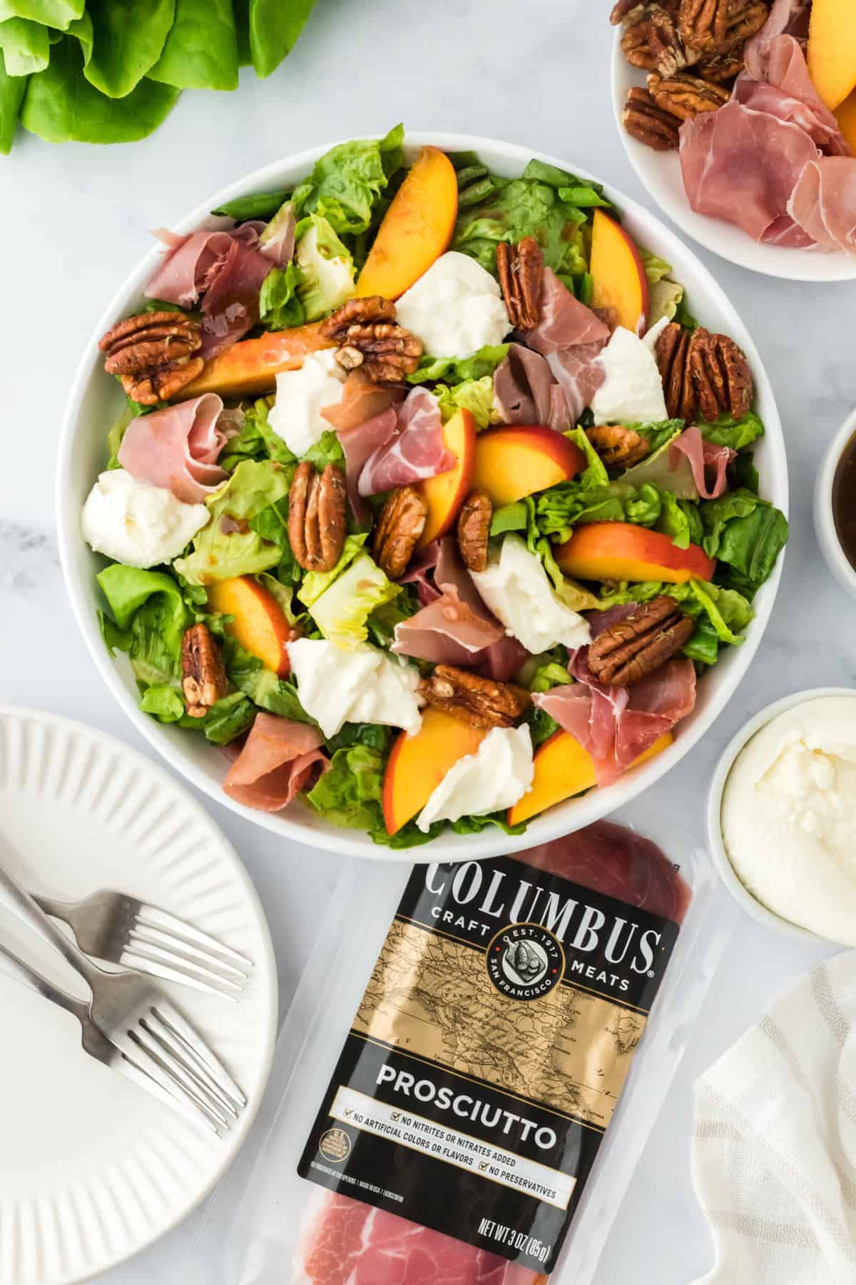 Peach burrata salad in a white bowl with Columbus meats prosciutto in a package next to it on a white background with some white plates and silver forks