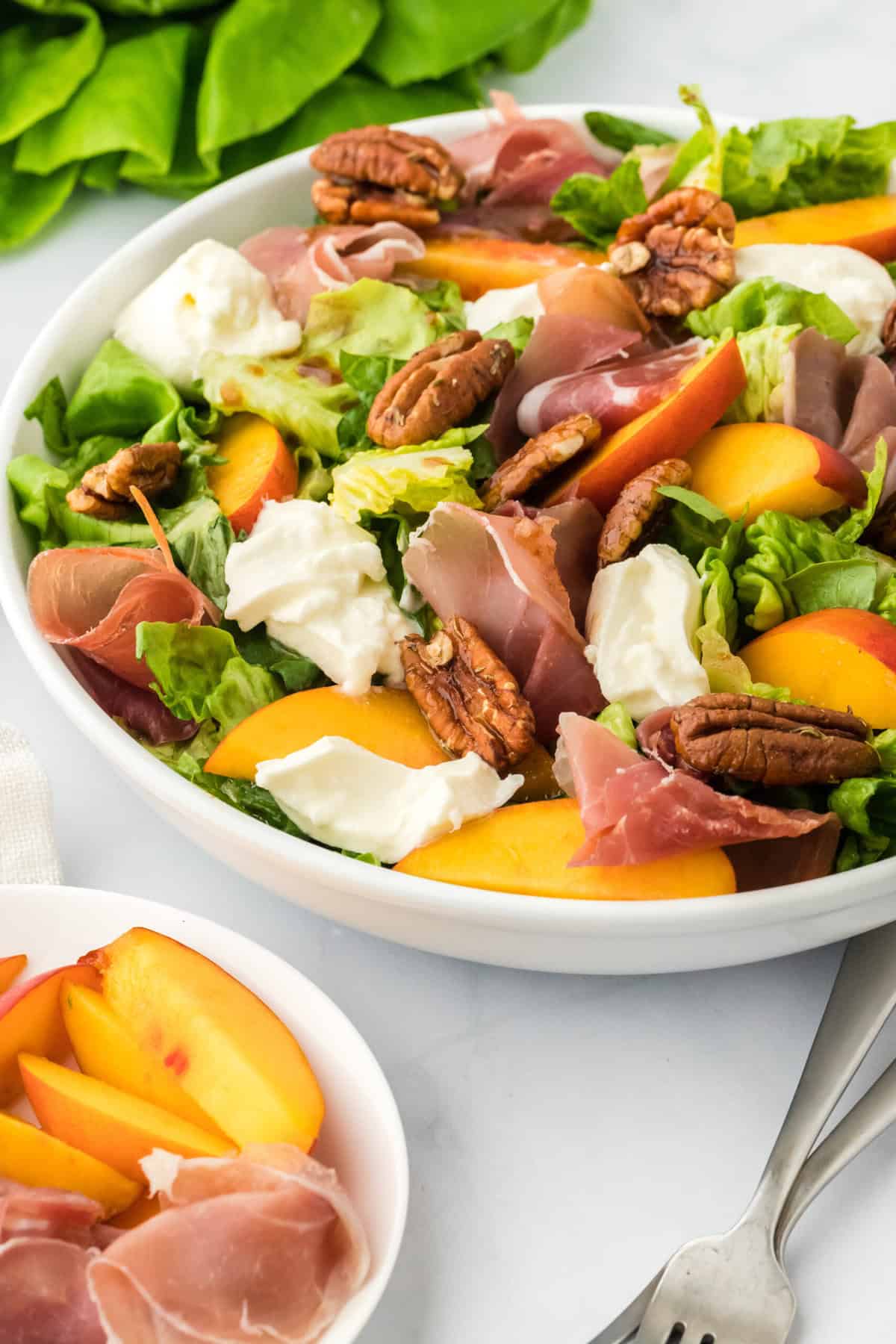 A large white bowl of peach salad recipe next to lettuce and peaches on a white plate on a white background